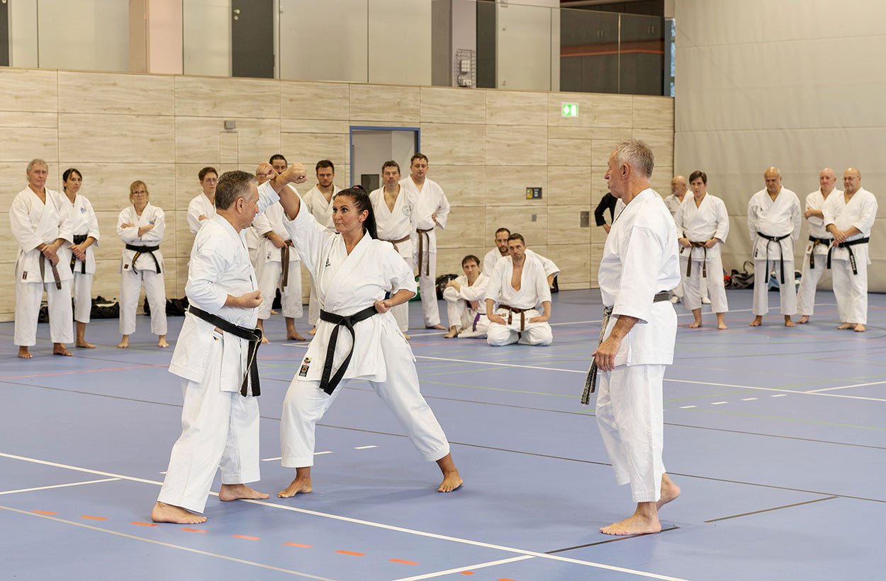 Eine Karateka führt eine Technik vor, während eine Gruppe im Halbkreis zusieht.